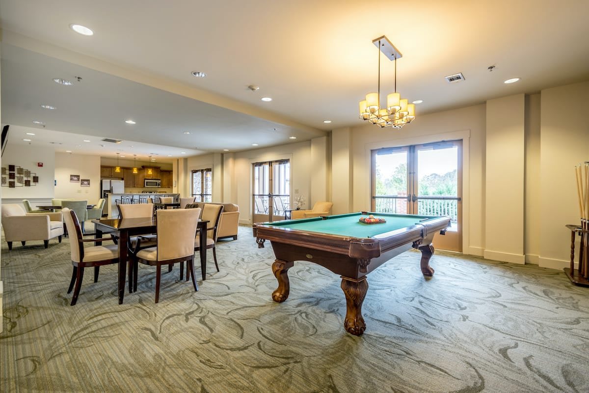 A spacious recreational room featuring a pool table and dining area, illuminated by natural light through large windows.