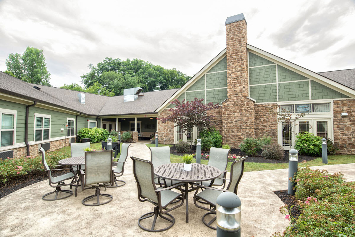 A cozy outdoor patio area with tables and chairs, set against a building with a stone facade and surrounded by lush landscaping.