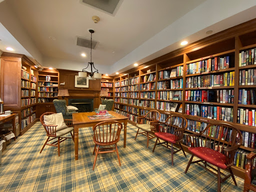 A cozy library featuring wooden bookshelves filled with books, a central table, and comfortable seating illuminated by warm lighting.
