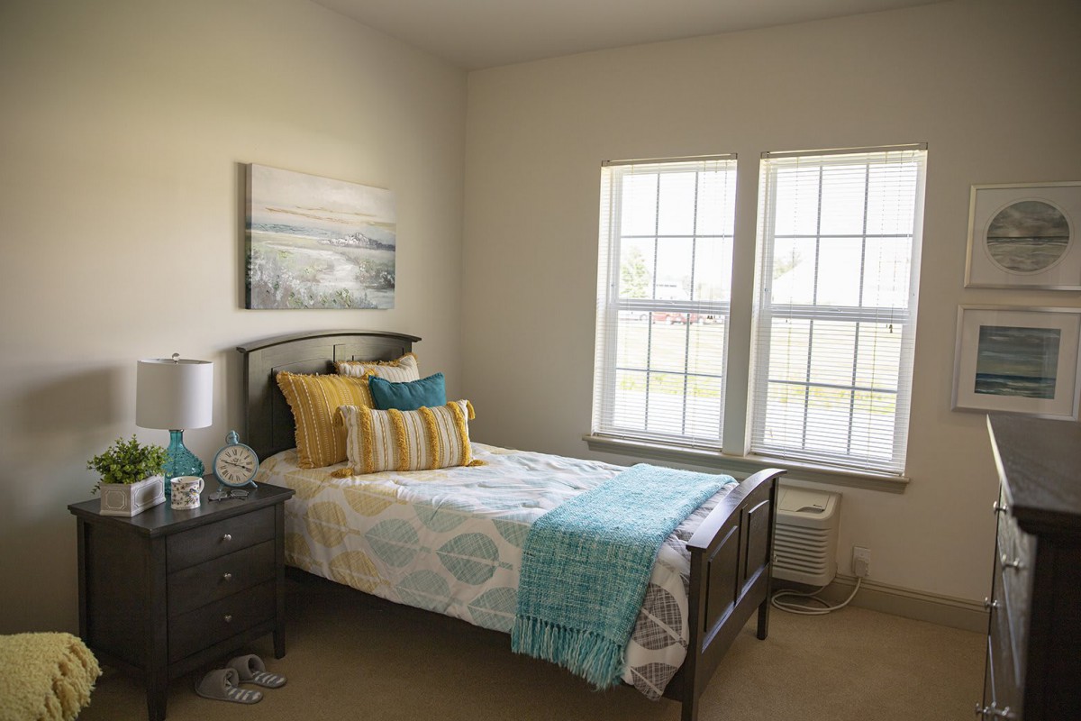 A cozy bedroom with a neatly made bed, colorful pillows, and large windows allowing natural light to fill the room.