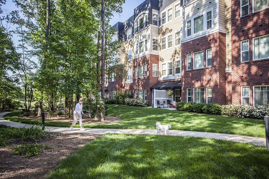 A person walks their dog along a pathway in front of a modern apartment building surrounded by trees and well-maintained lawns.