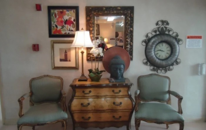 A vintage-style interior with an antique dresser, decorative mirror, framed art pieces, a table lamp, and two elegant chairs.