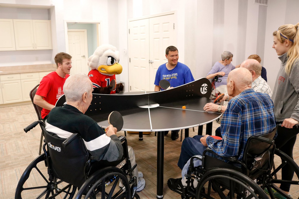 A group of elderly individuals in wheelchairs enjoy a game of table tennis with the help of staff and a mascot in a nursing home setting.