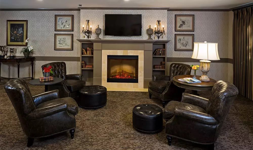 A cozy living room with leather chairs and a central fireplace, featuring decorative lamps and a television above the mantel.