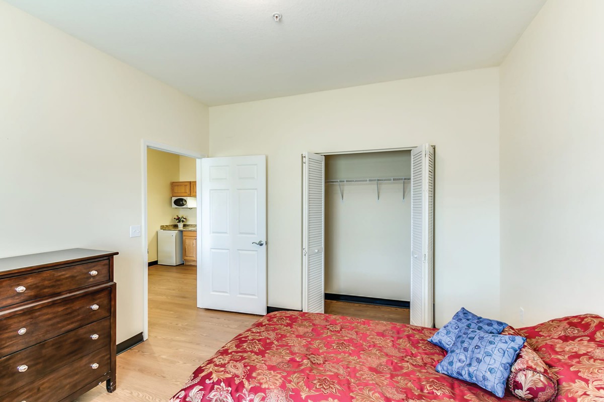 A bedroom with an open closet, bed with red bedding, and a doorway leading to another room.