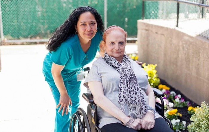 A nurse poses with a patient in a wheelchair outside on a sunny day.