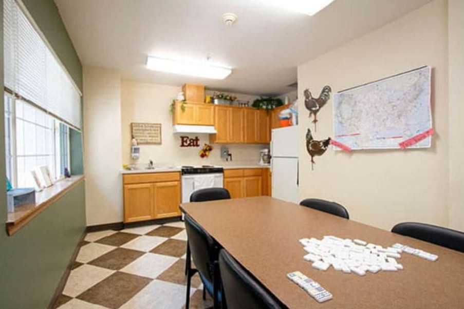 A small break room kitchen with a table and chairs, featuring cabinets, a refrigerator, and decorative wall art.