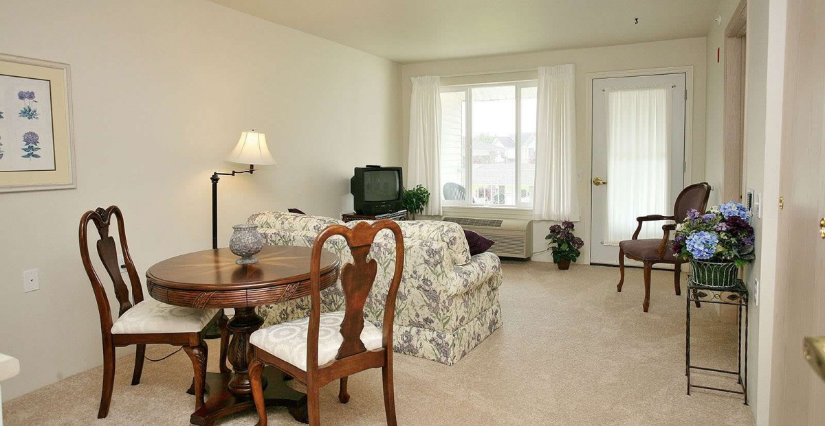 A cozy living room featuring a wooden table with chairs, a plush sofa, a television, and a standing lamp by the window.