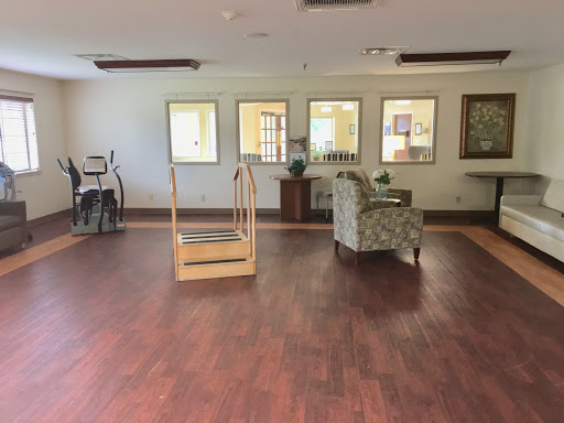 A spacious and empty lounge area featuring a wooden floor, a patterned chair, and rehabilitation equipment in the corner.