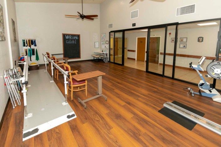 A rehabilitation room equipped with physical therapy exercise tools and chairs, featuring a wooden floor and large glass doors.