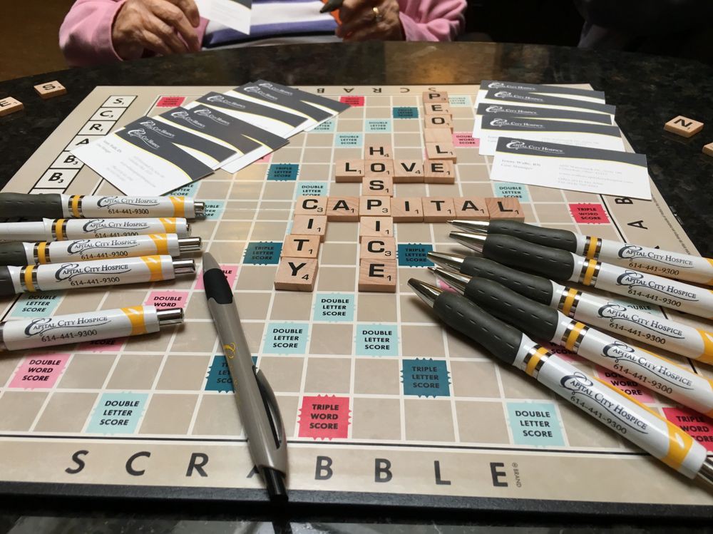 A Scrabble board with tiles forming words, surrounded by pens and a person holding a book.