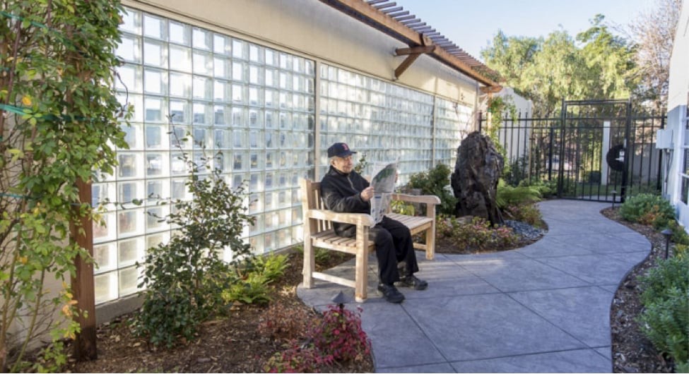 A person sitting on a bench in a garden, reading a newspaper.