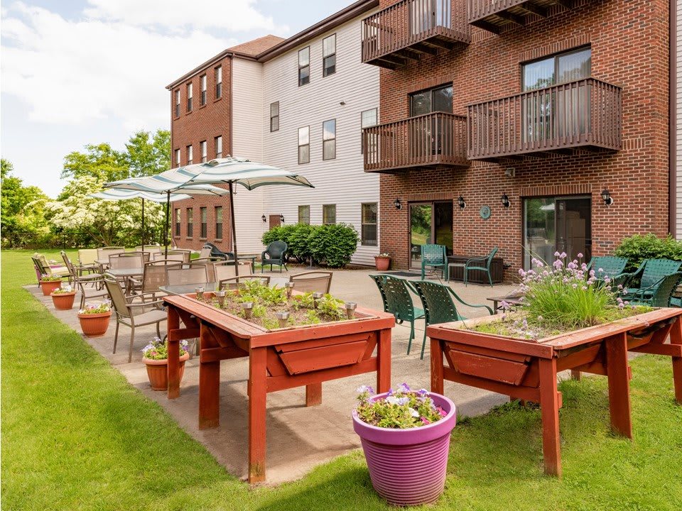 An outdoor garden area with raised planters, tables, and chairs under umbrellas beside a multi-story building.