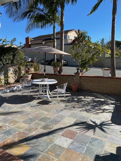 A sunny outdoor patio area with a table, chairs, and an umbrella surrounded by potted plants and palm trees.
