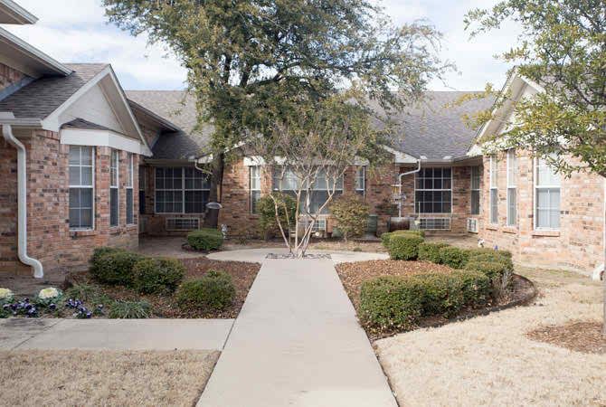 A brick residential house with landscaped bushes and trees, featuring a concrete sidewalk leading to the entrance.