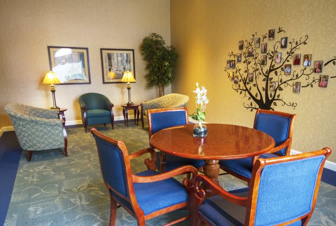 A cozy lobby seating area with a round wooden table surrounded by blue upholstered chairs, decorative wall art, and a potted plant.