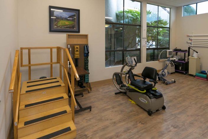 A well-lit gym with exercise equipment including a recumbent bike and stairs for rehabilitation exercises.