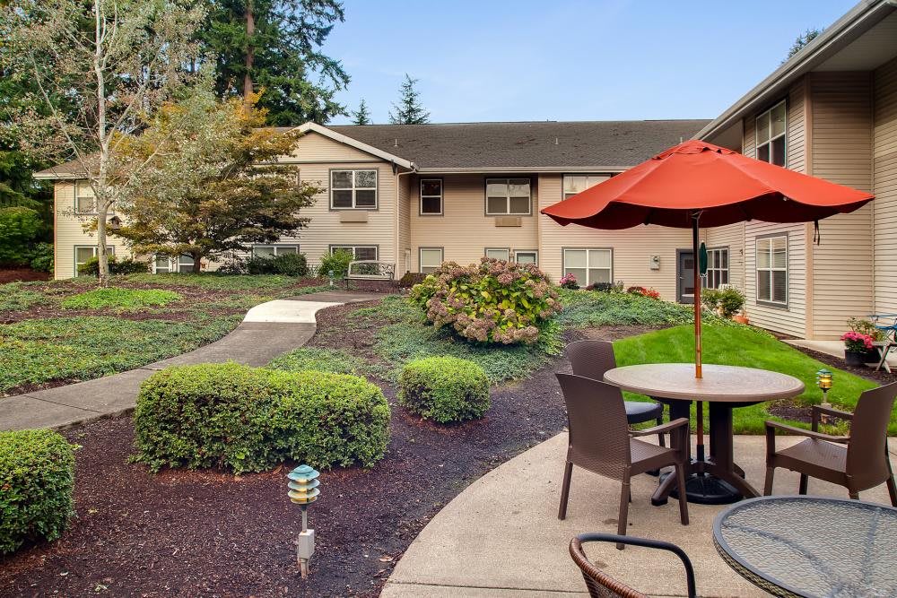 A serene outdoor patio area with tables, chairs, and a red umbrella surrounded by well-maintained landscaping.