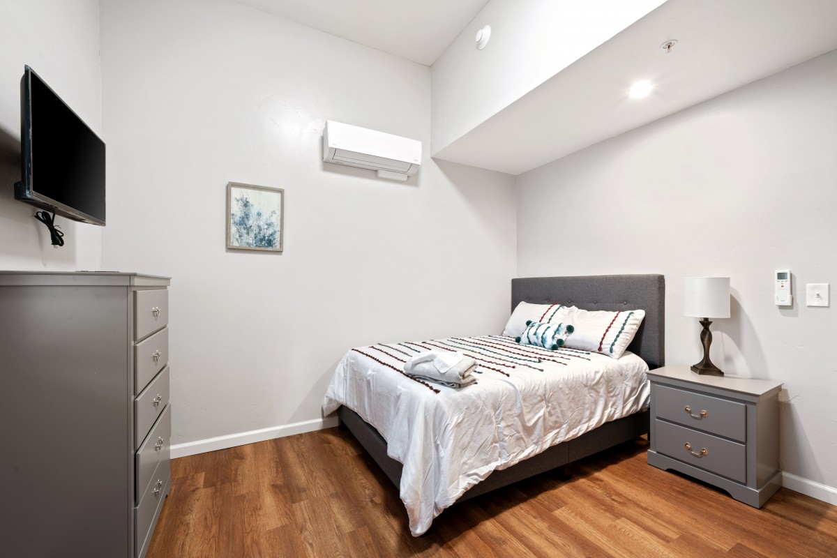 A modern and minimalist bedroom featuring a comfortable bed, wooden flooring, and an air conditioning unit.