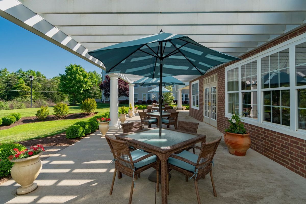 A sunny patio with outdoor seating and umbrellas under a pergola, surrounded by a garden.