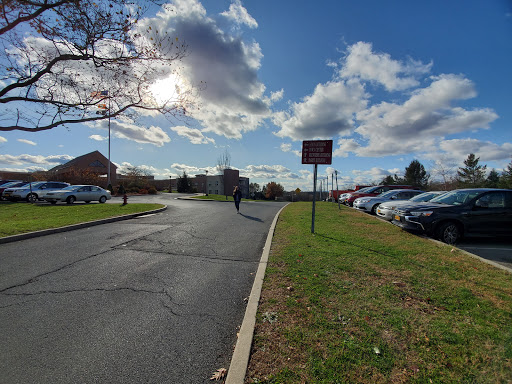 A lone figure walks along a winding road beside a parking lot under a partly cloudy sky.