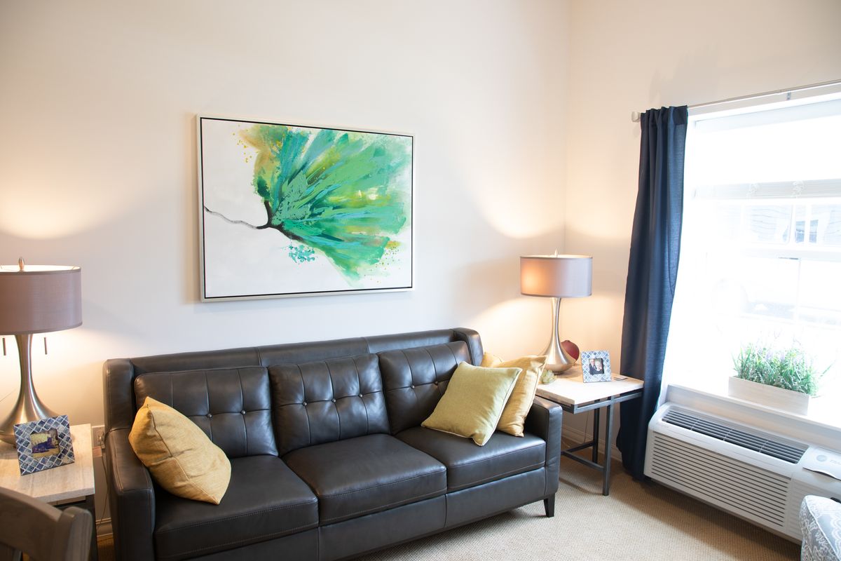 A modern living room with a dark leather sofa, colorful artwork, and a cozy lamp by the window.