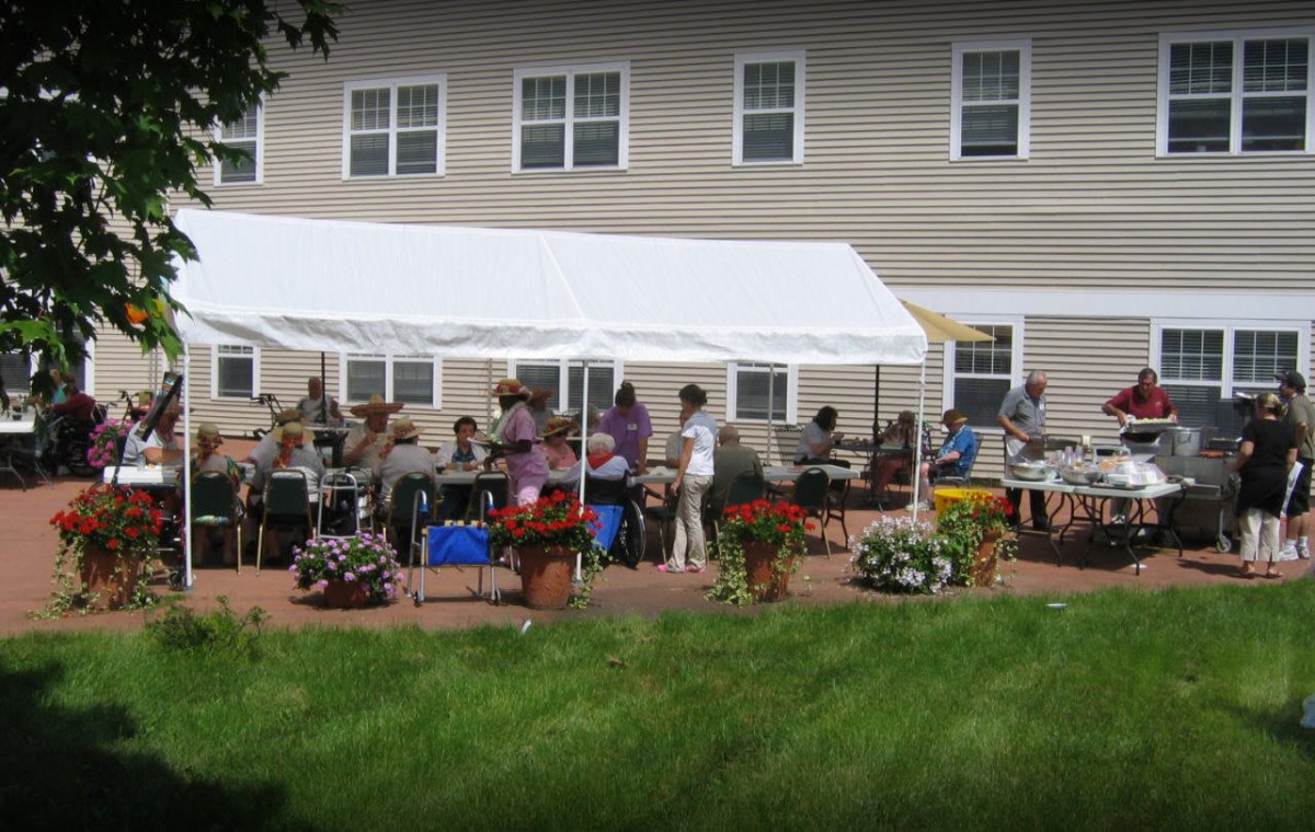 A group of people gathered under a white tent outside a house with a grassy lawn and flower arrangements.