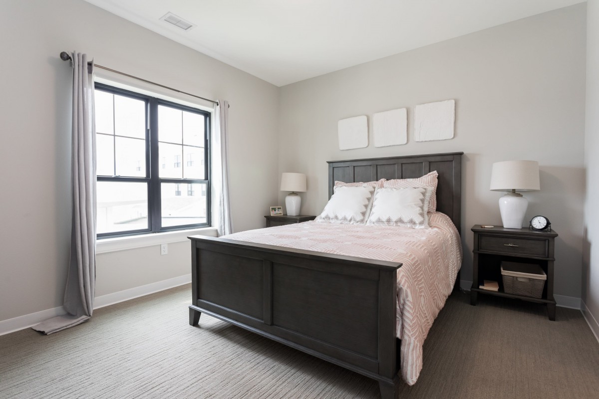A modern bedroom with a large bed, gray walls, and natural light from a window.