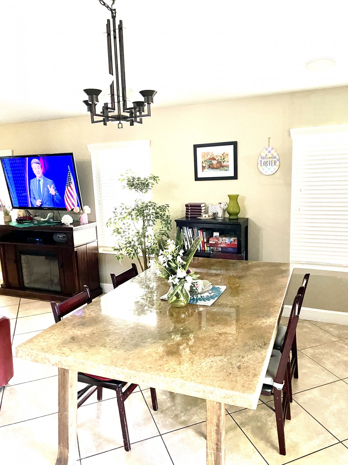 A well-lit dining room with a large table, chairs, and a TV on in the background.