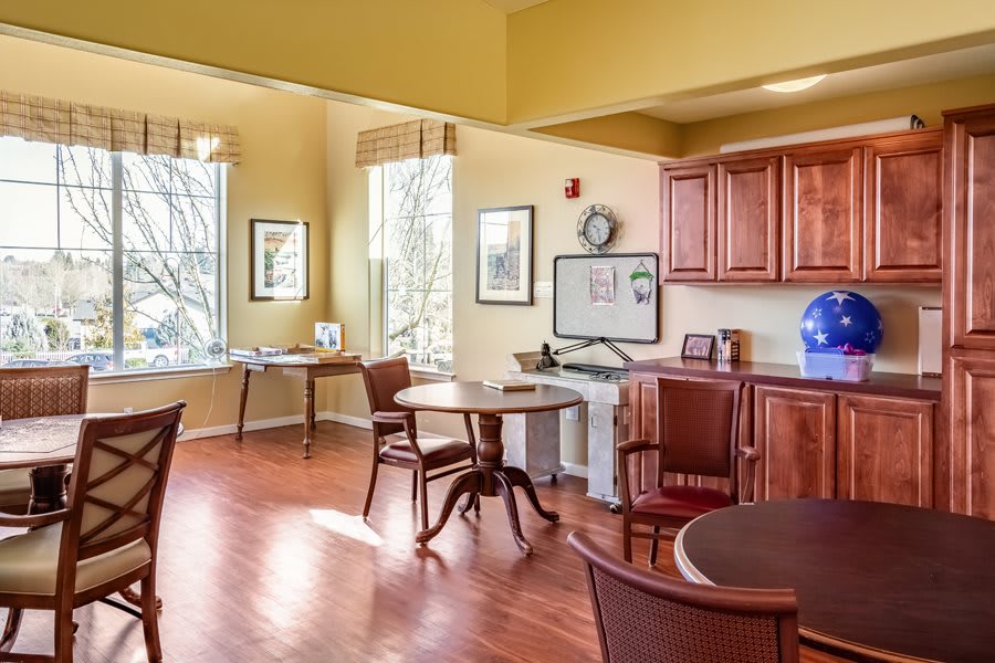 A cozy dining area with wooden tables and chairs, featuring warm wood flooring and natural light from large windows.
