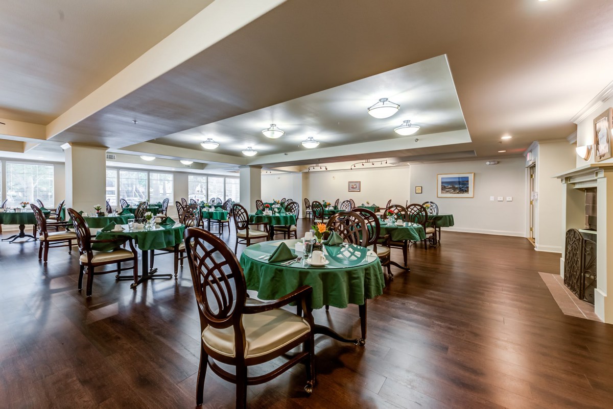 A spacious dining room with green tablecloths and wooden chairs arranged neatly on a wooden floor.