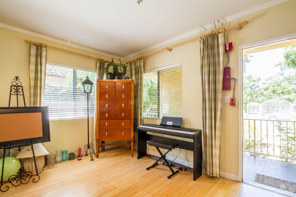 A cozy living room featuring a digital piano and a wooden chest of drawers against a bright, windowed wall with light curtains.