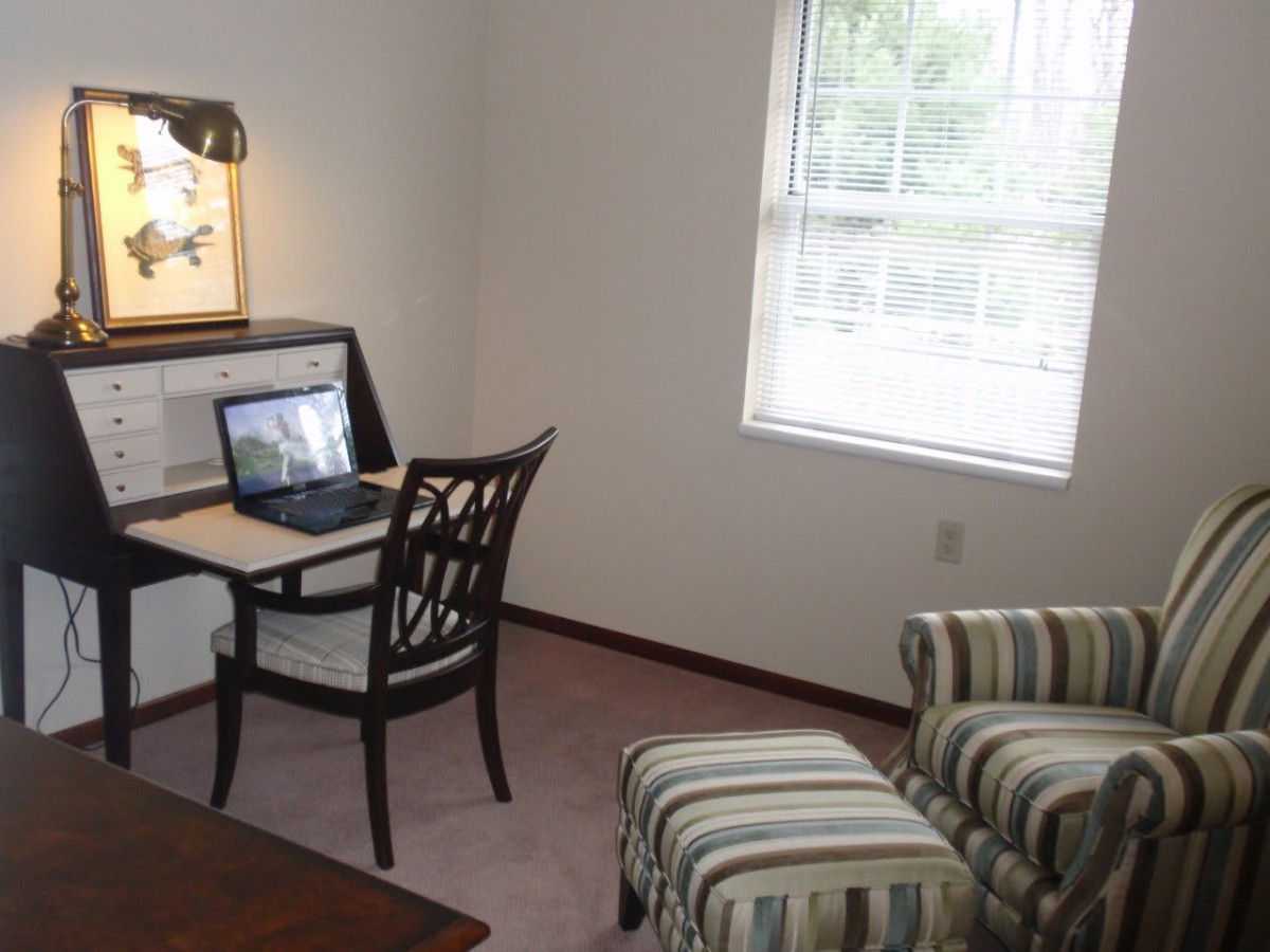 A cozy home office with a desk, chair, laptop, window, armchair, and ottoman.