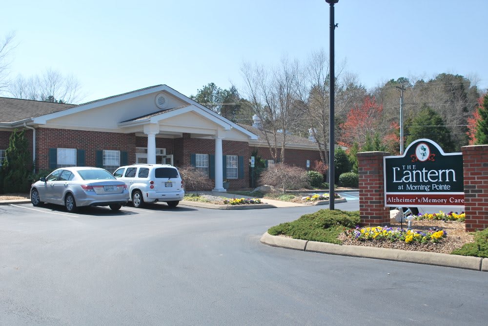 A small brick building with a parking lot and cars in front, surrounded by trees.