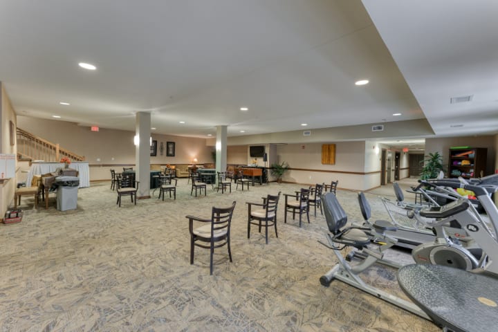 A spacious indoor fitness center with exercise equipment, chairs, and tables on a carpeted floor.