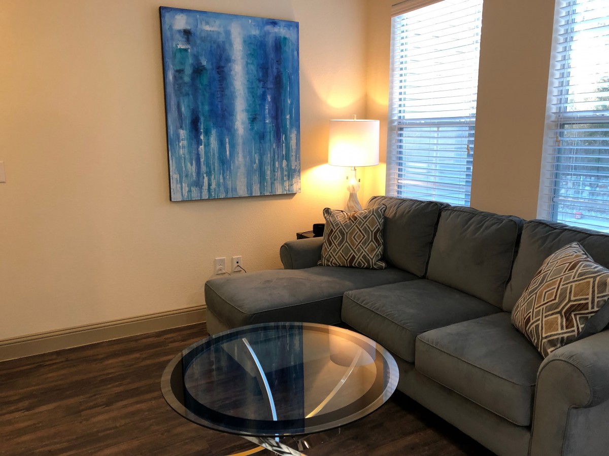 A stylish living room with a gray sectional sofa, glass coffee table, and blue abstract painting on the wall.