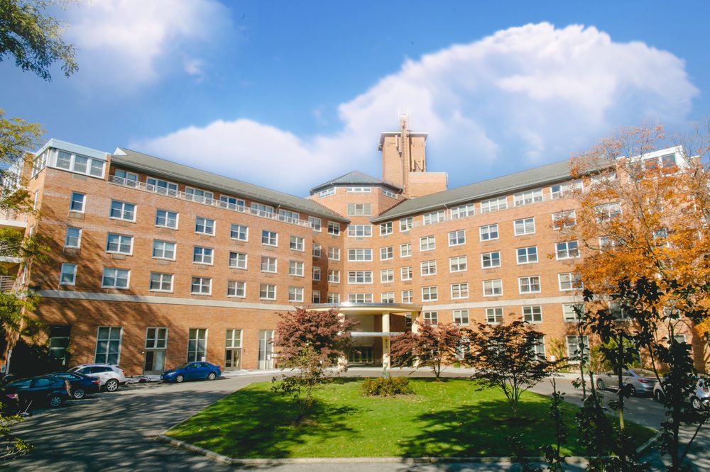 A large brick hospital building with a clear blue sky and scattered clouds in the background.
