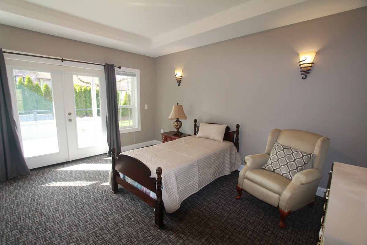A cozy bedroom with a single bed and armchair, featuring a carpeted floor and natural light from a window and door.