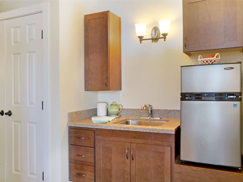 A small kitchen area featuring wooden cabinets, a sink, a countertop, and a compact fridge under warm lighting.