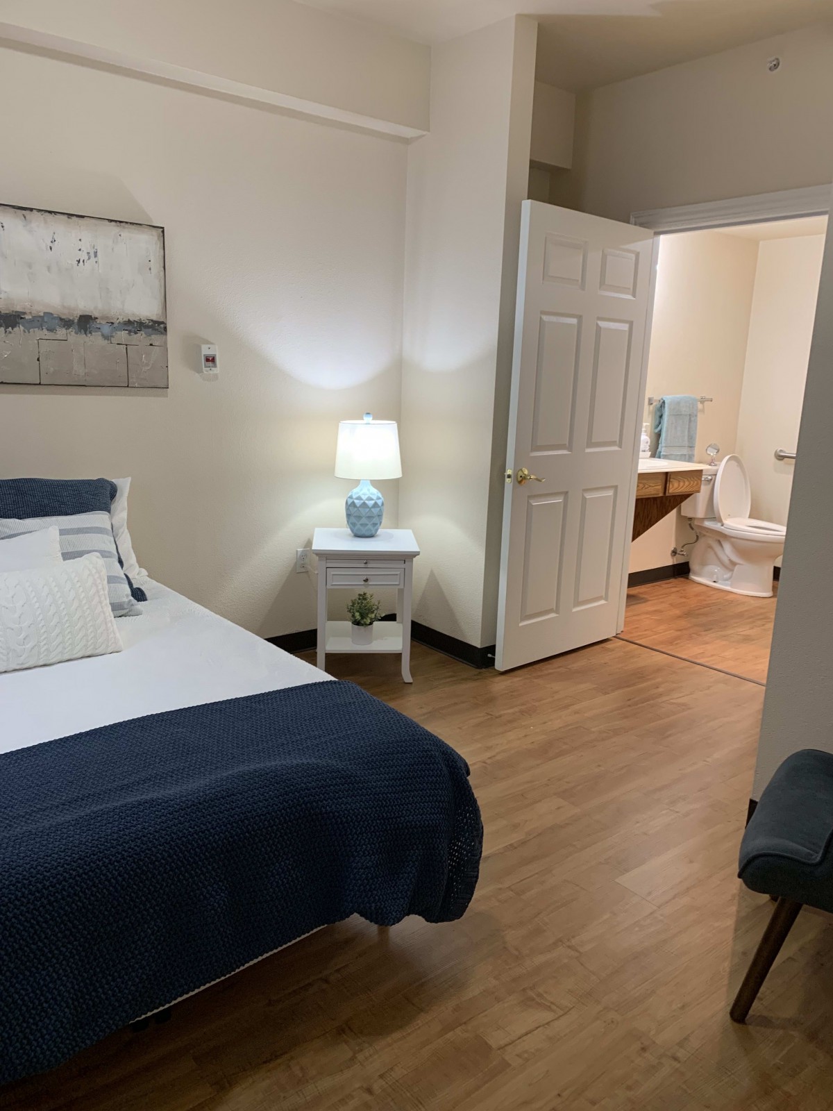 A cozy bedroom with a neatly made bed, nightstand with a lamp, and an open doorway leading to a bathroom.