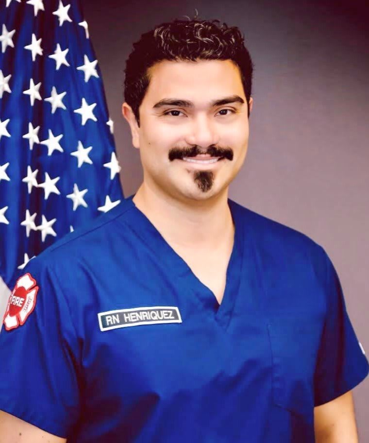 A man in blue medical scrubs stands in front of an American flag.