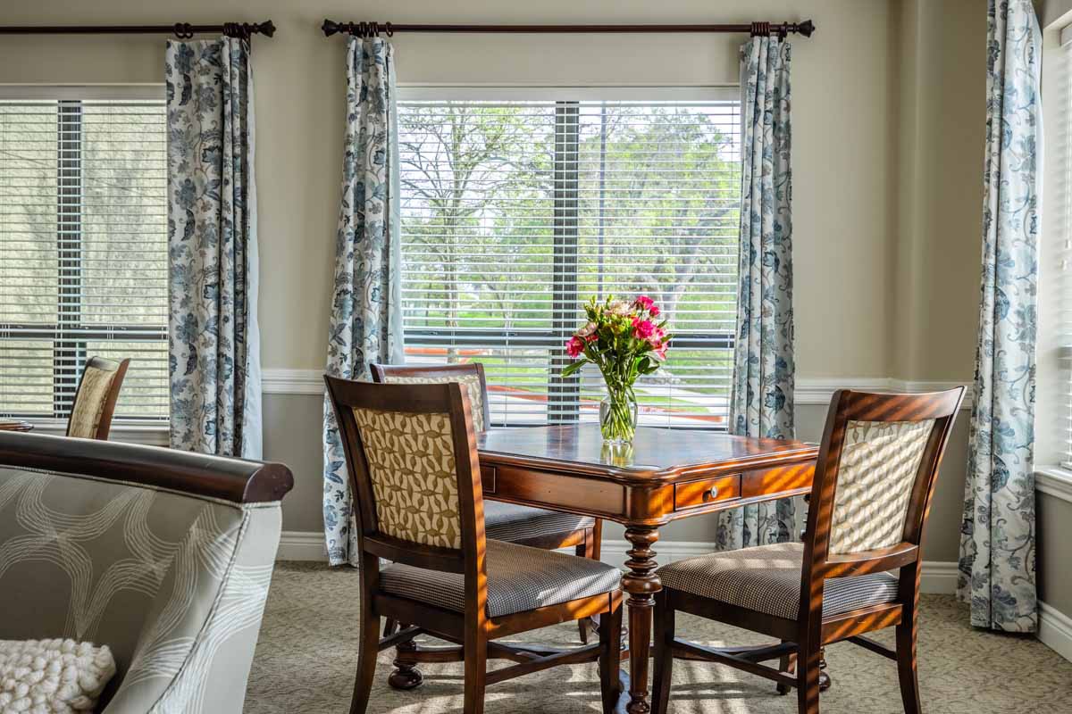 A cozy dining room with a wooden table and chairs, adorned by a vase of flowers near a large window.