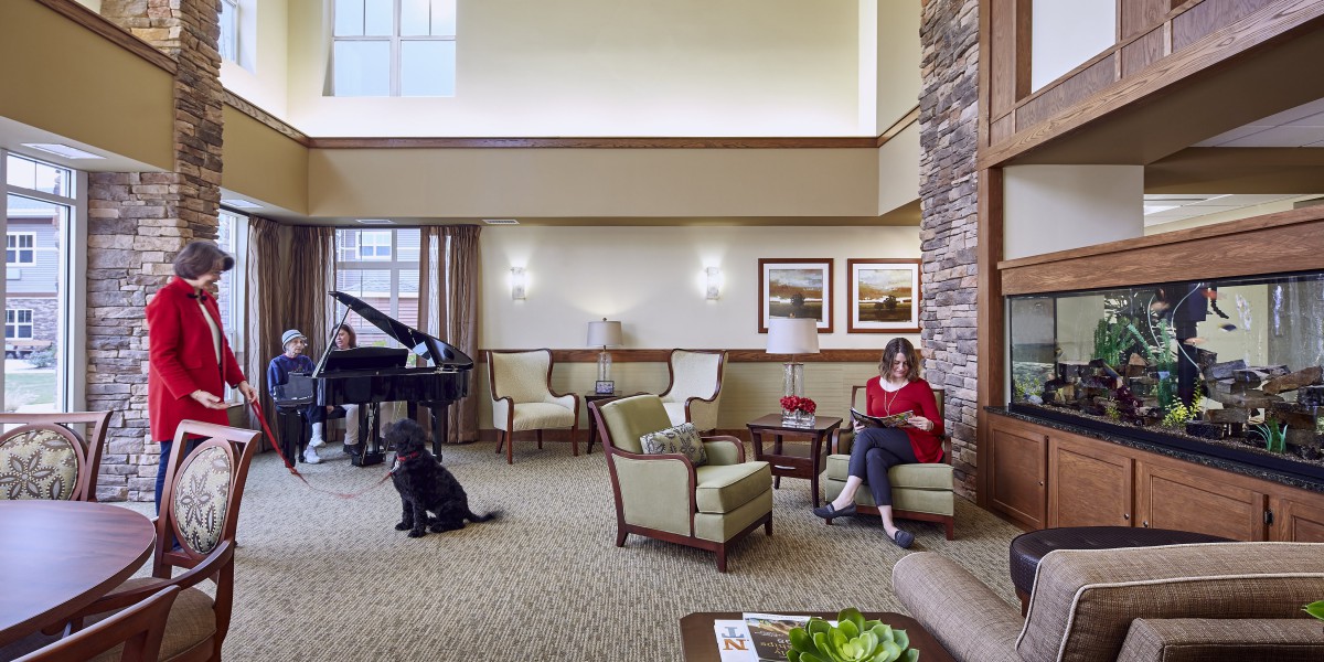 A cozy living room featuring a grand piano, a woman reading on a sofa, and a dog sitting on the carpet.