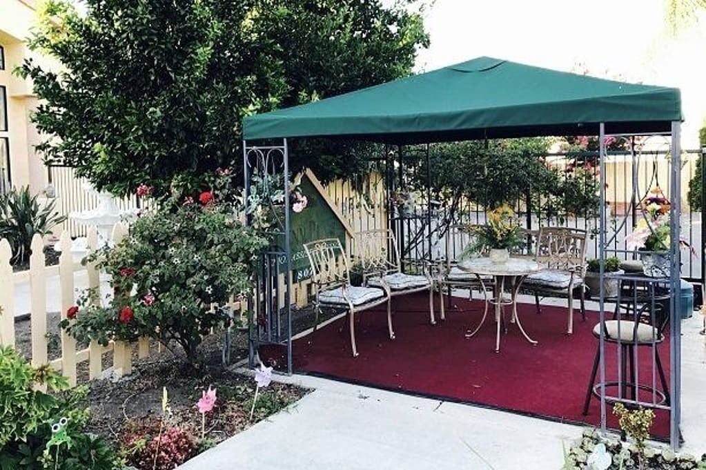 A cozy outdoor gazebo with patio chairs and a table, surrounded by lush greenery and flowers.