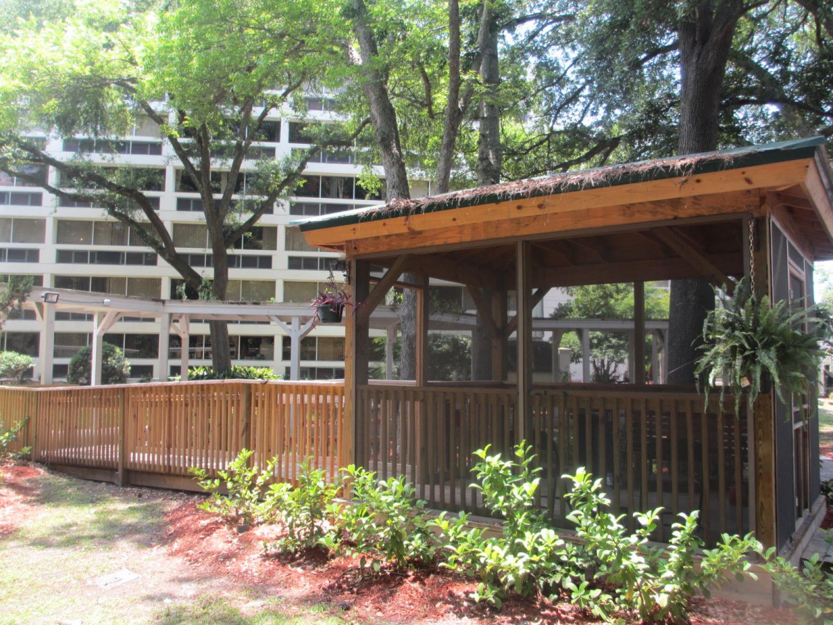 A wooden gazebo surrounded by lush greenery, with a modern building in the background.