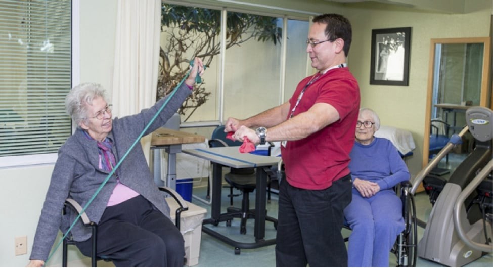A healthcare professional assists an elderly person with exercises using resistance bands in a rehabilitation setting.