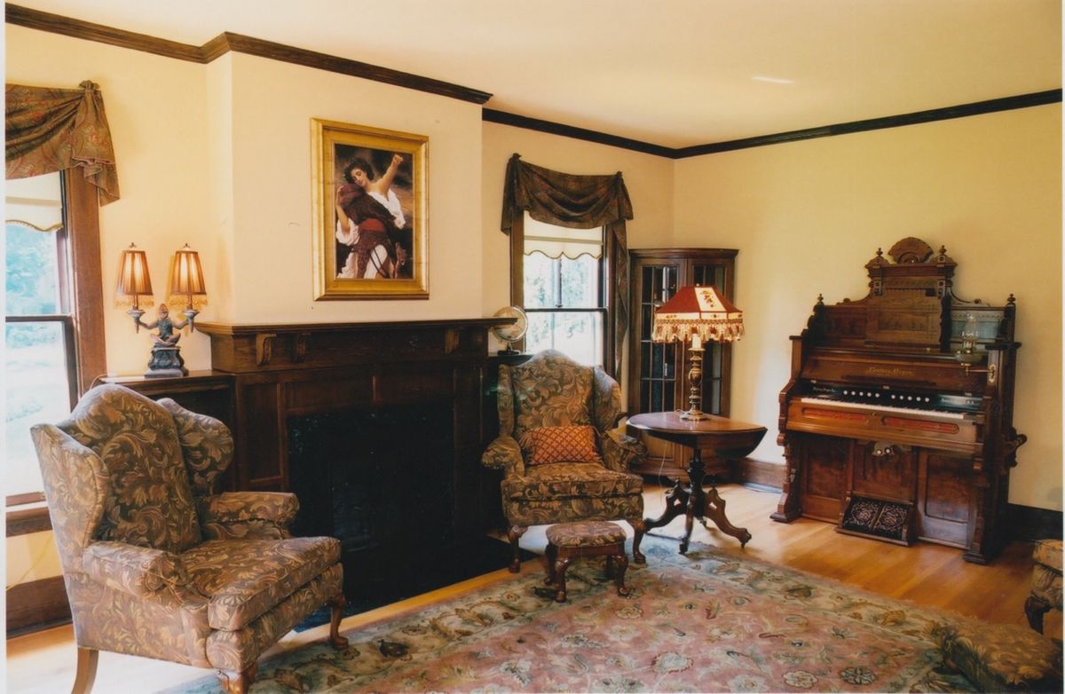 A cozy vintage-style living room featuring a fireplace, patterned armchairs, and an antique organ.