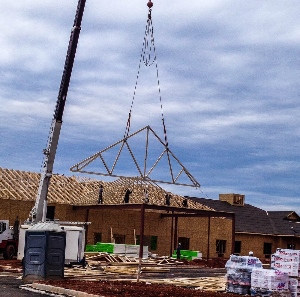 A crane lifts a roof truss for installation at a construction site under a cloudy sky.