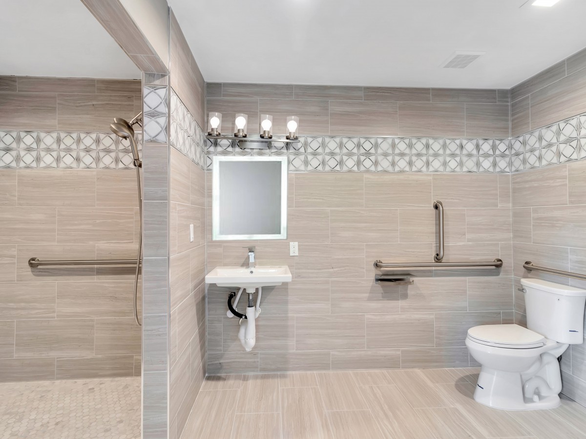A modern bathroom with tiled walls, featuring a sink with a mirror, toilet, and safety handrails.
