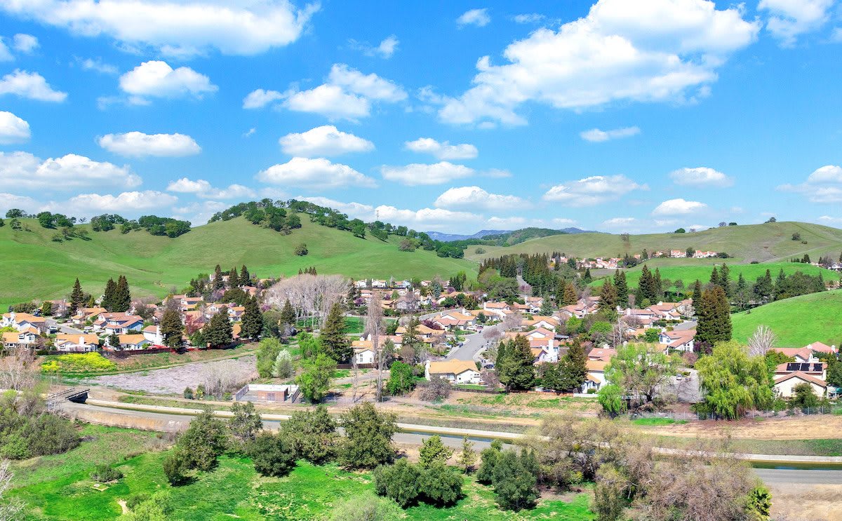 A scenic view of a lush green landscape with rolling hills and a quaint village under a bright blue sky dotted with clouds.